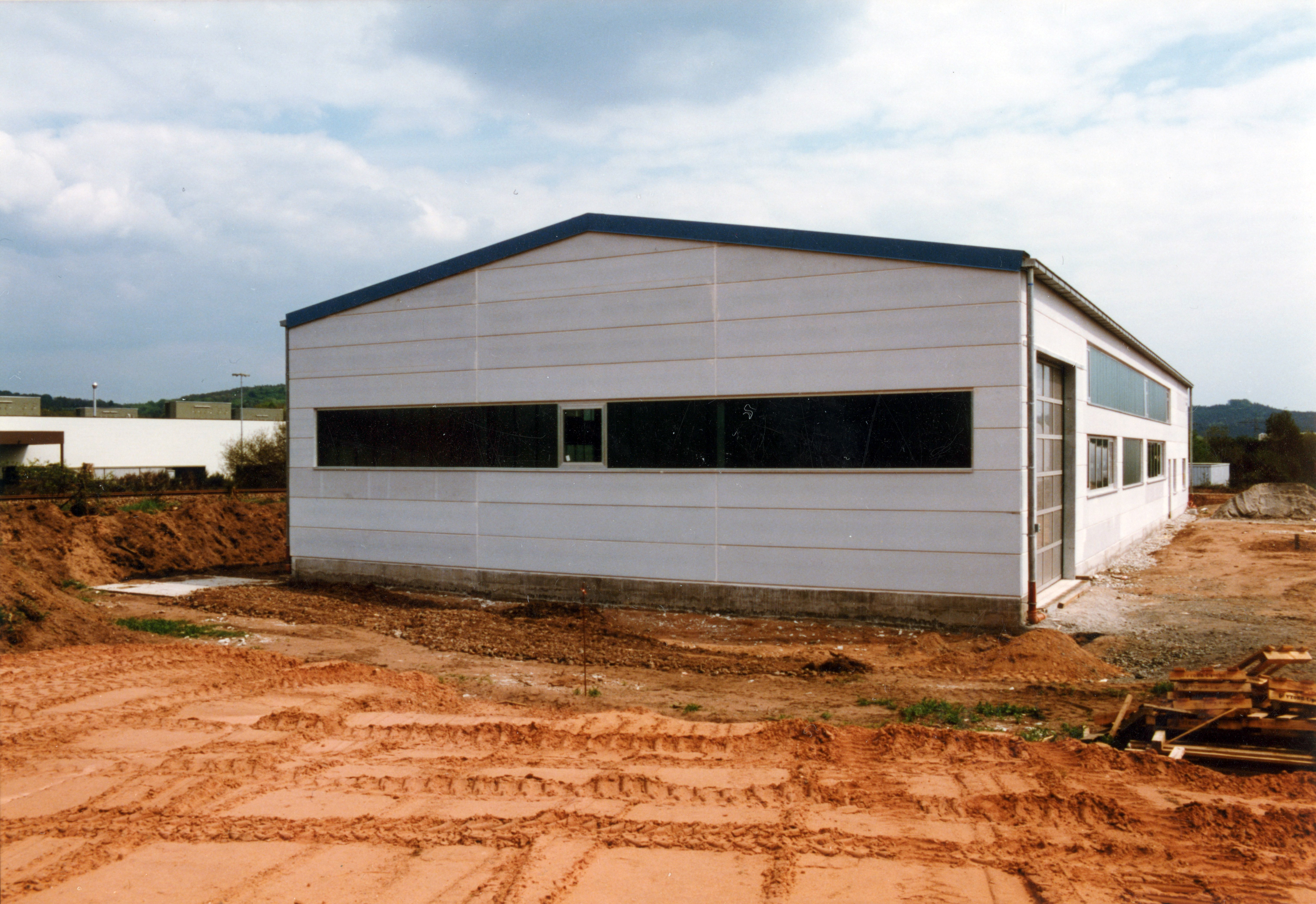 Lagerhalle von der Seite. Die Halle sieht fertig aus, die Erde drumherum ist karg mit Baggerspuren.