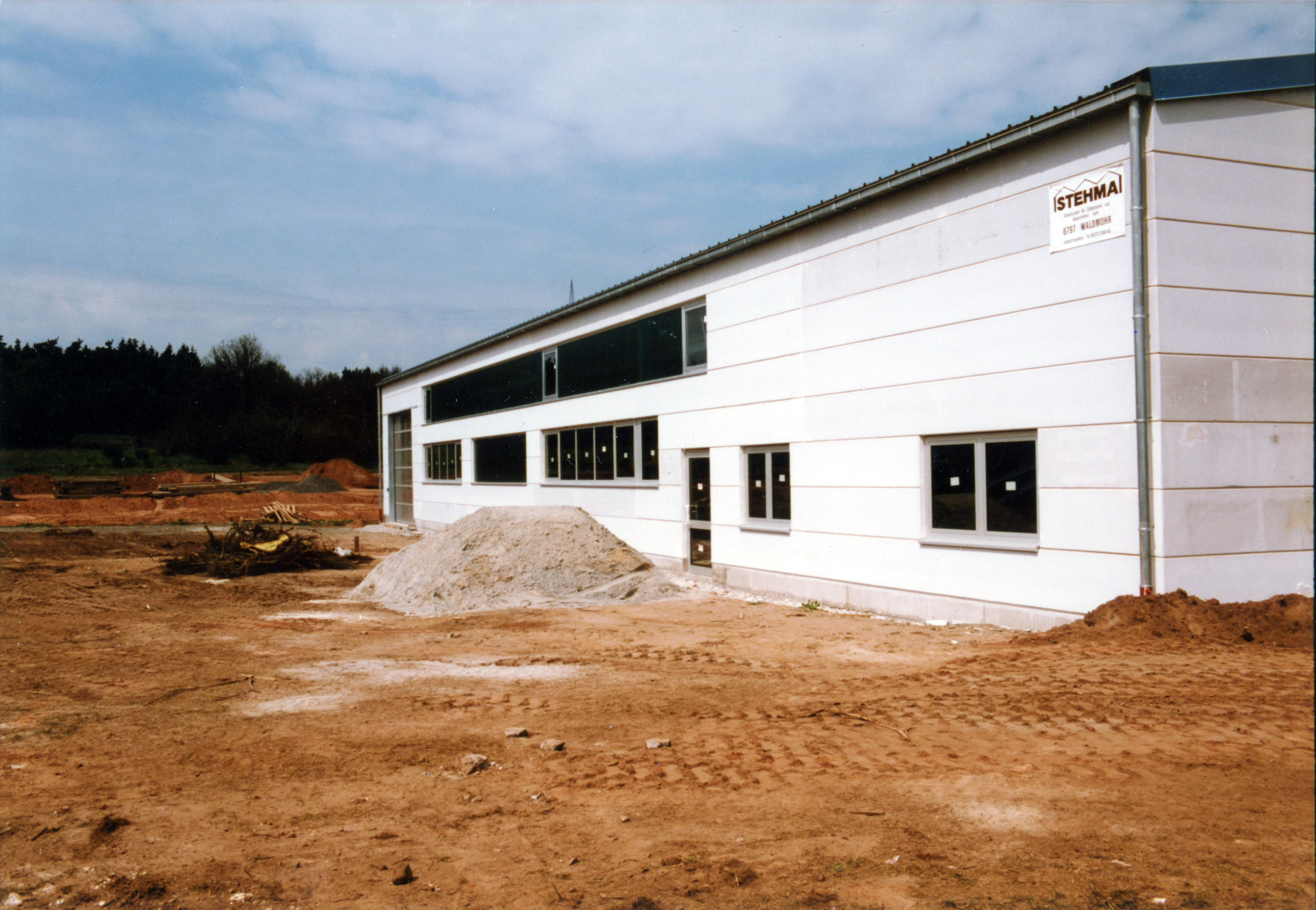Lagerhalle von vorne. Die Halle sieht fertig aus, die Erde drumherum ist noch karg mit Baggerspuren.
